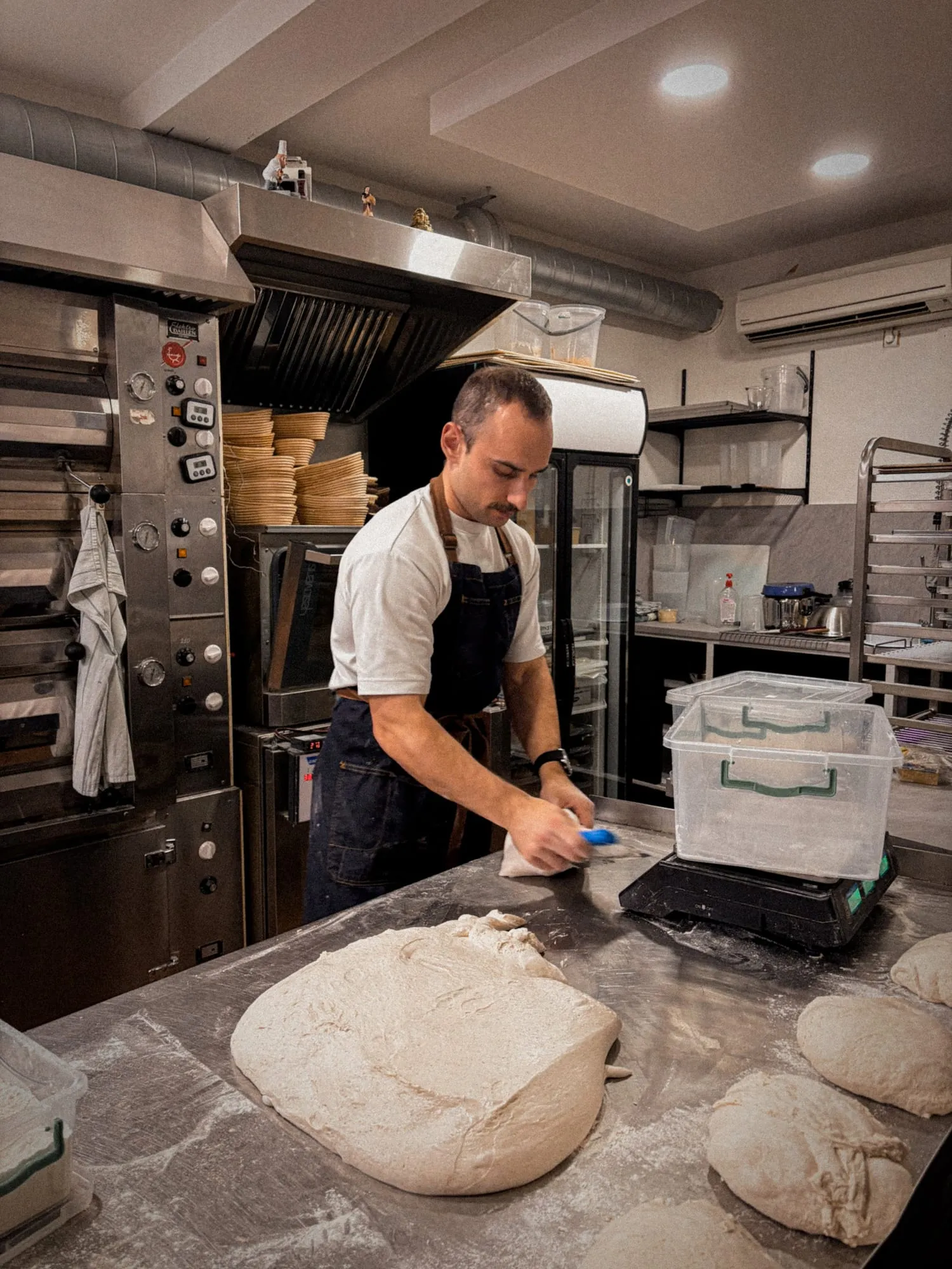 The baker dividing sourdough on a floured bench, proofing baskets stacked by the deck oven behind him